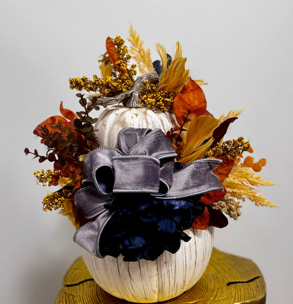 Decorative pumpkin with autumn leaves and a large bow on a wooden stump against a white wall.
