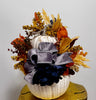 Decorative pumpkin with autumn leaves and a large bow on a wooden stump against a white wall.
